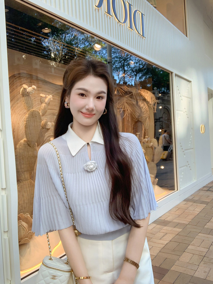 Woman wearing a light blue pleated blouse with a white collar and a white skirt, carrying a small quilted chain bag