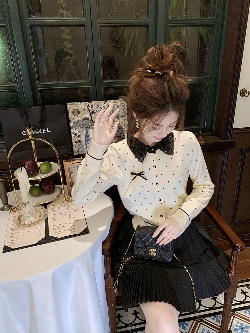 Woman wearing a cream polka dot blouse with black collar and a black pleated skirt, holding a small black quilted handbag