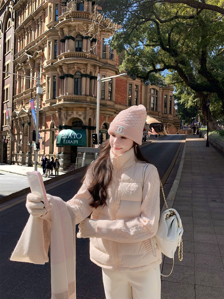 Woman wearing a pale pink winter outfit with a knit beanie, puffer vest, gloves, scarf, and white backpack on a city street