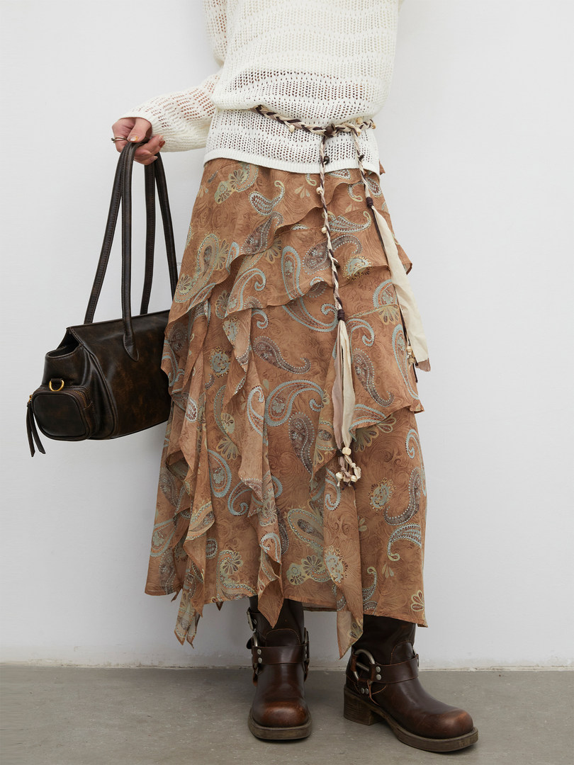 Model wearing a layered paisley midi skirt with a white knit sweater and brown boots, holding a dark handbag