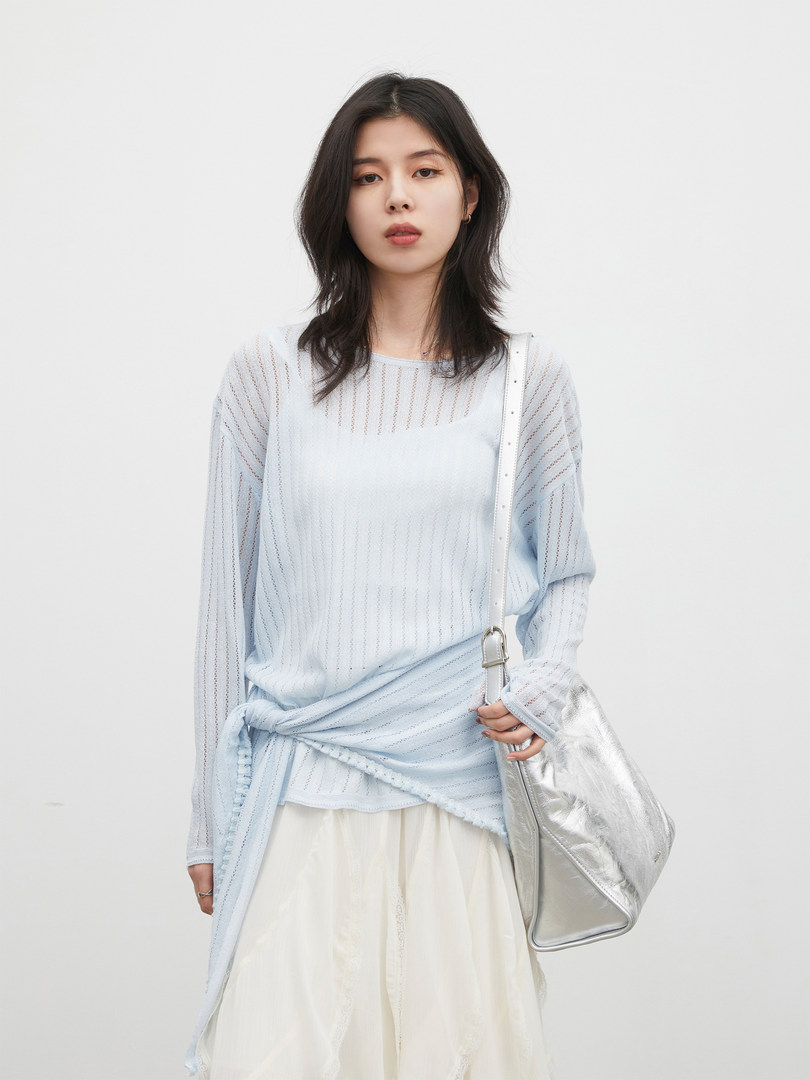 Model wearing a light blue sheer long-sleeve top with a white skirt and silver shoulder bag