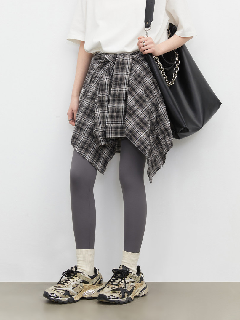 Model wearing a white top, plaid wrap skirt, gray leggings, sneakers, and a black shoulder bag