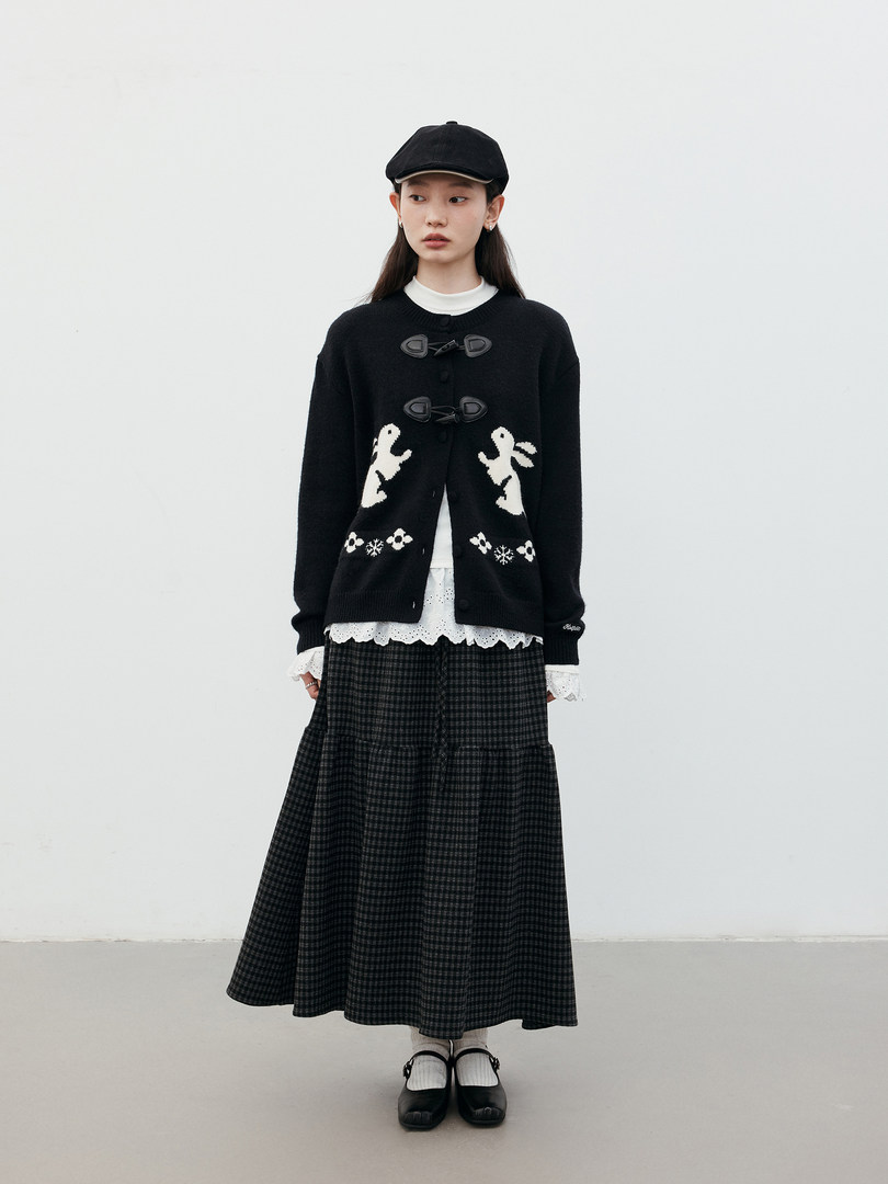 Model wearing a black cardigan with a plaid midi skirt and black cap