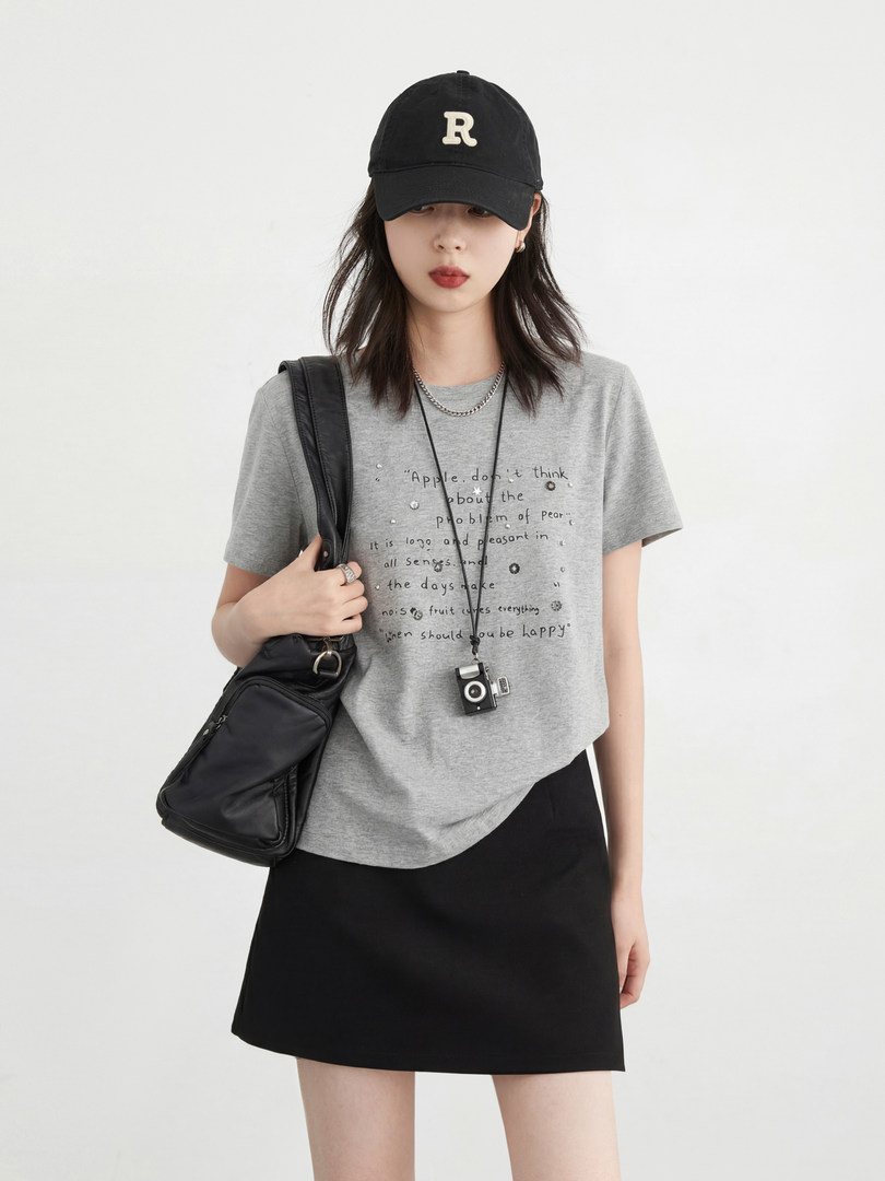 Model wearing a gray graphic T-shirt with black skirt, cap, and shoulder bag