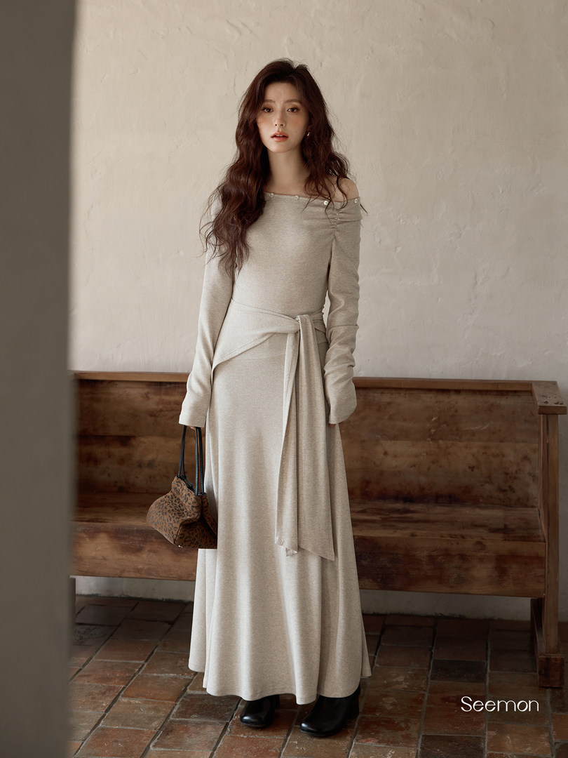 Model wearing a beige off-shoulder long-sleeve maxi dress with a tie waist and carrying a patterned handbag