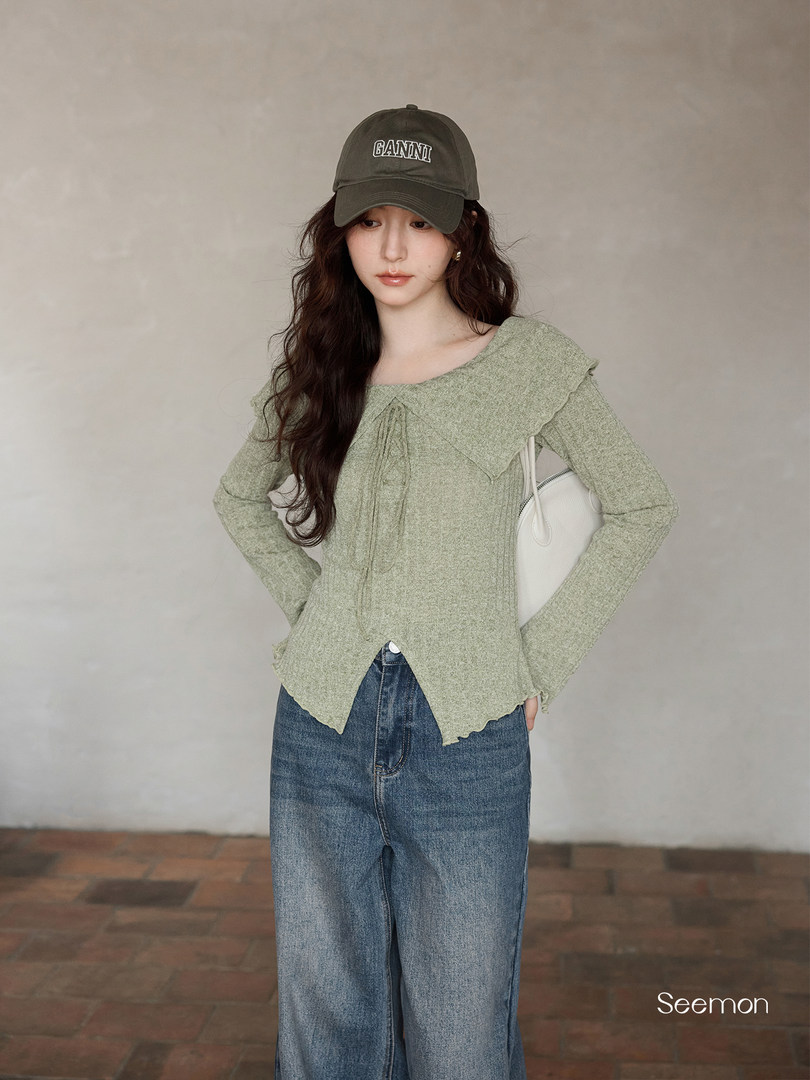 Model wearing a light green knit top with a large collar, blue jeans, and a dark cap