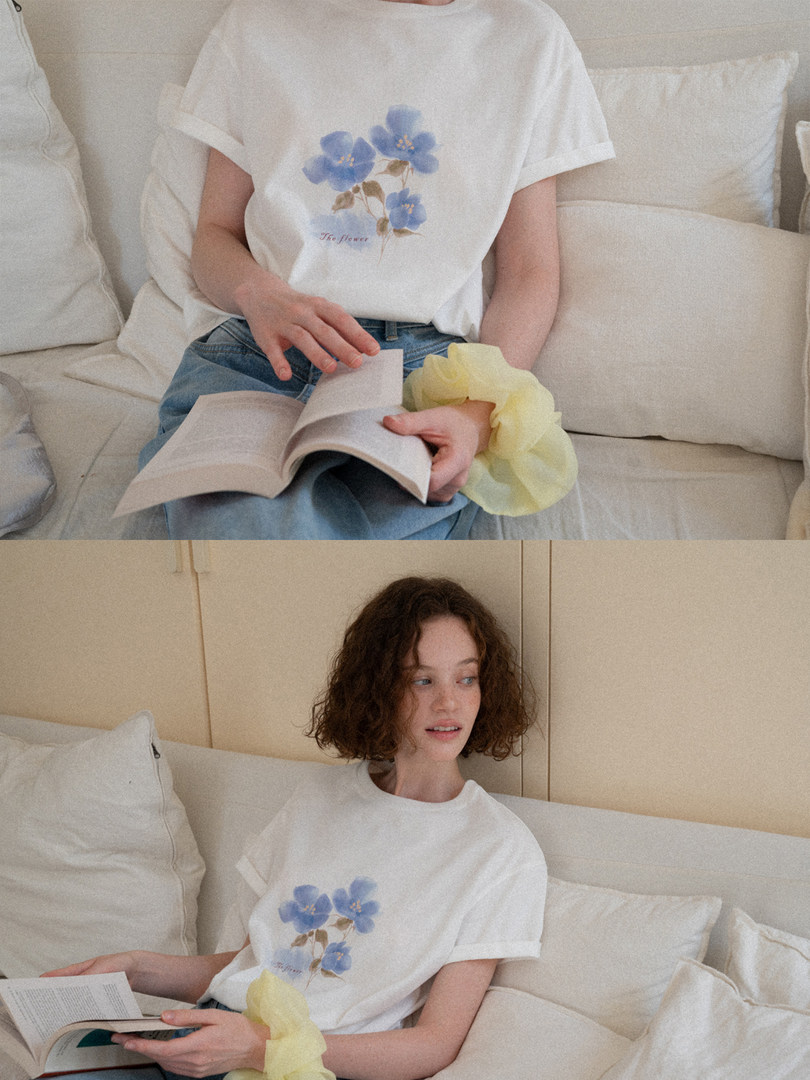 White graphic T-shirt with blue floral print worn by a model sitting on a couch
