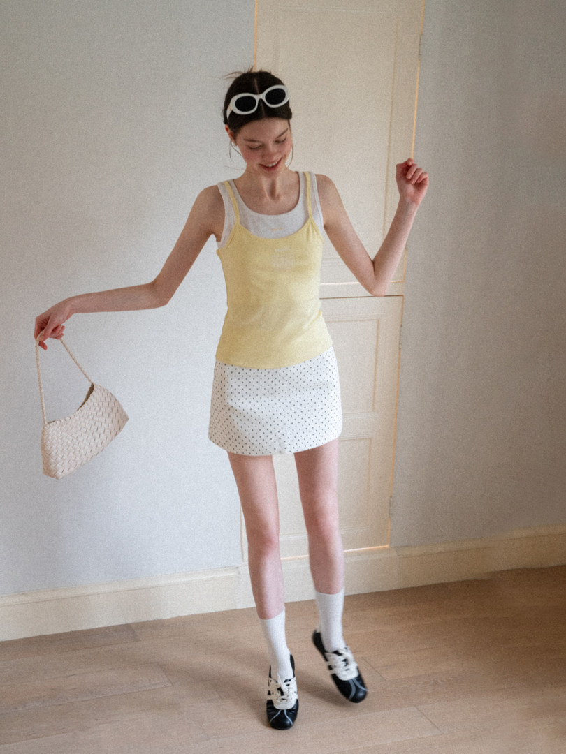 Model wearing a yellow tank top, white mini skirt, and black shoes while holding a woven handbag