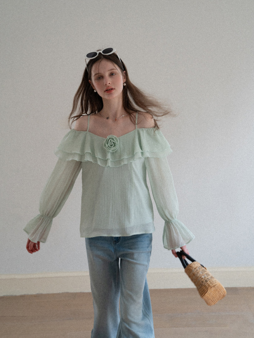 Model wearing a mint off-shoulder ruffled blouse with long sheer sleeves