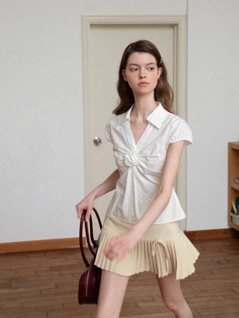 Model wearing a white short-sleeve blouse with a cream pleated mini skirt and handbag