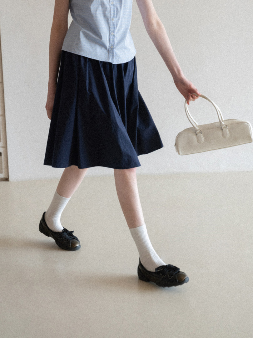 Model wearing a light blue sleeveless top with a navy skirt and holding a cream handbag