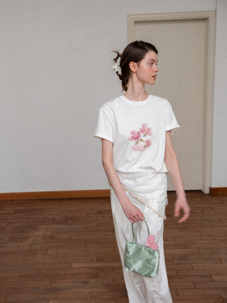 Woman wearing a white floral graphic T-shirt and white pants, holding a small green bag