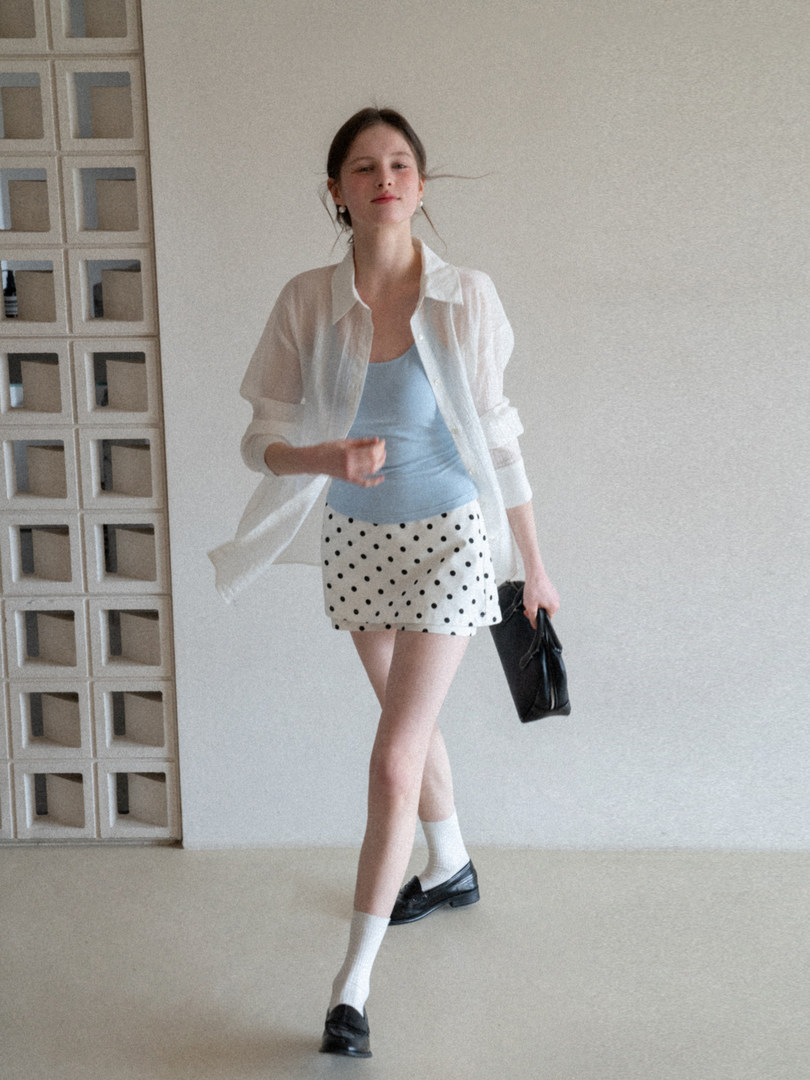 Model wearing a sheer white shirt over a blue tank top with a polka dot mini skirt and black loafers