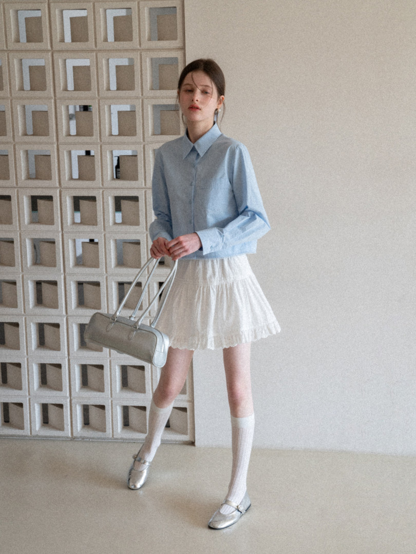 Model wearing a light blue button-up shirt with a white pleated skirt and silver flats, holding a silver handbag.