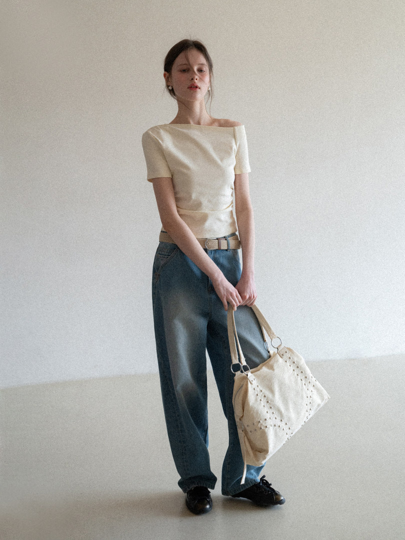 Model wearing a cream off-shoulder top with relaxed blue jeans and carrying a beige handbag