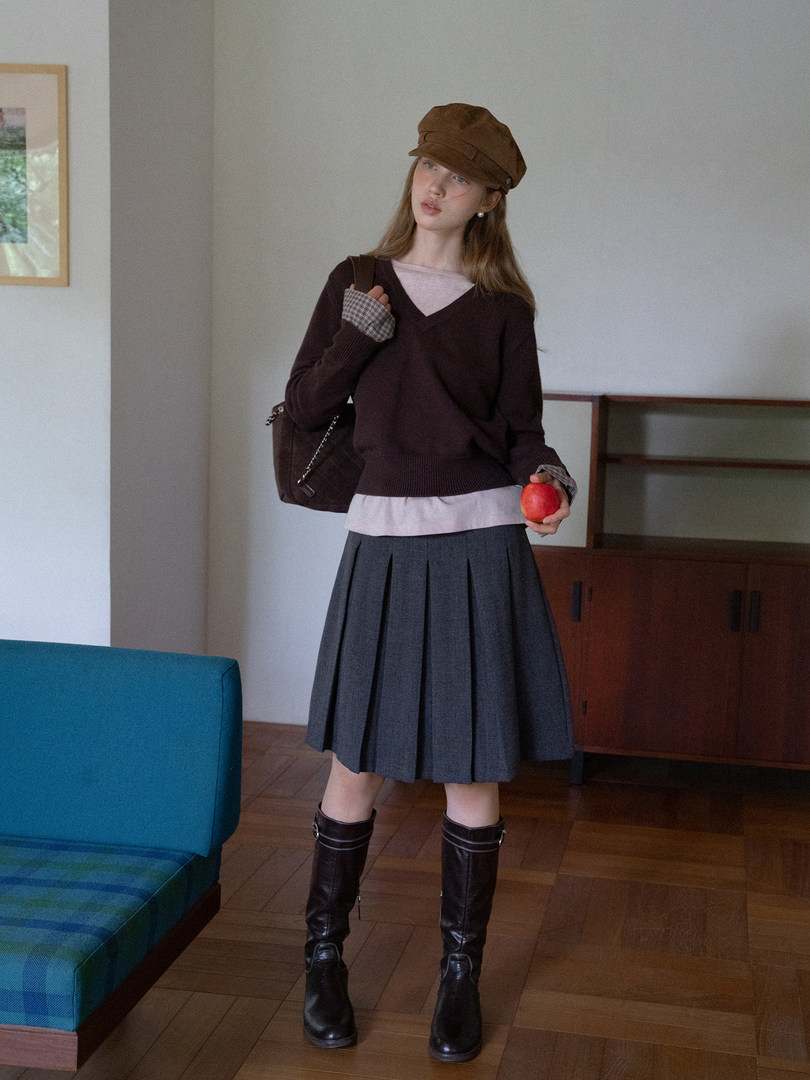 Model wearing a dark sweater, pleated skirt, brown hat, and knee-high boots indoors