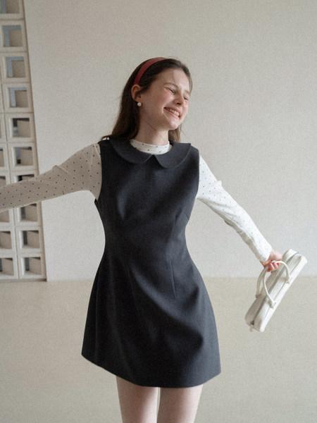Model wearing a black sleeveless pinafore dress over a white polka dot long-sleeve top