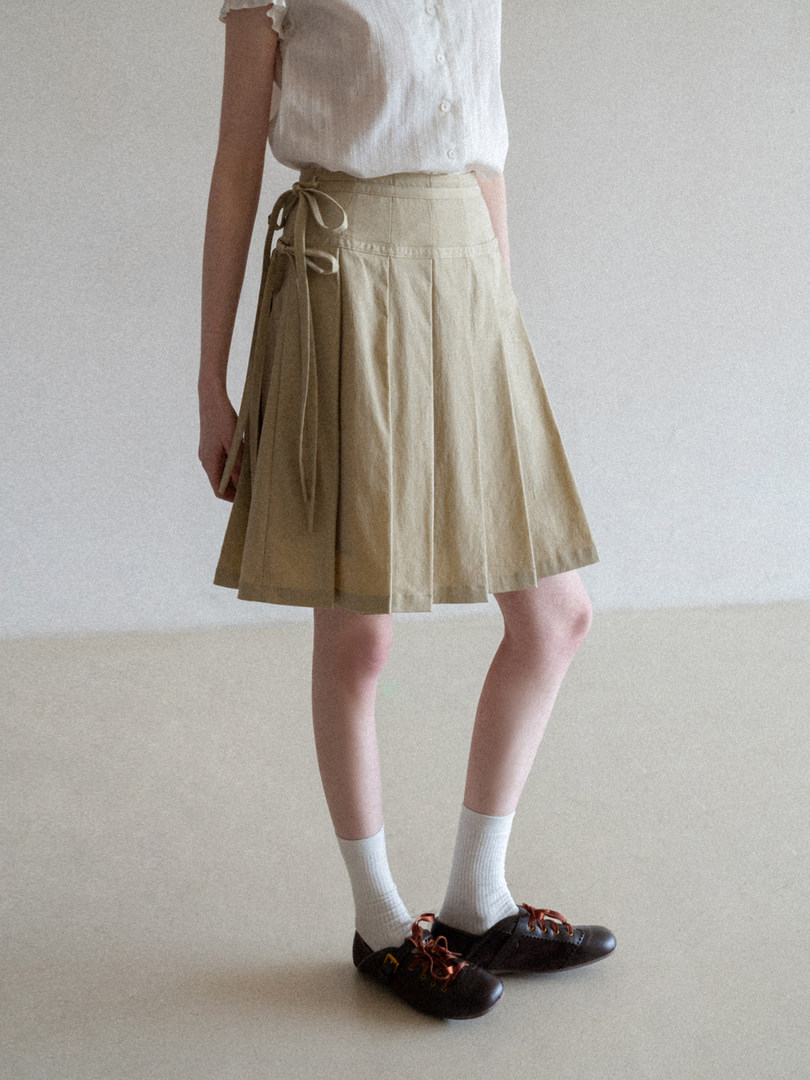 Model wearing a beige pleated wrap skirt with a white blouse and black shoes