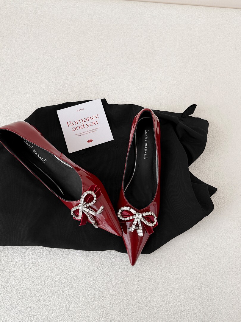 Pair of glossy red pointed-toe flats with rhinestone bow details on a black cloth background