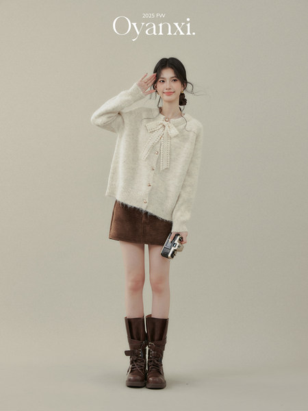 Female model wearing a cream fuzzy cardigan with bow detail, brown mini skirt, and tall lace-up boots