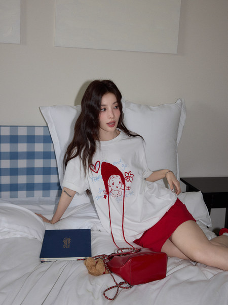 Model wearing a white graphic T-shirt with red shorts and holding a red handbag on a bed