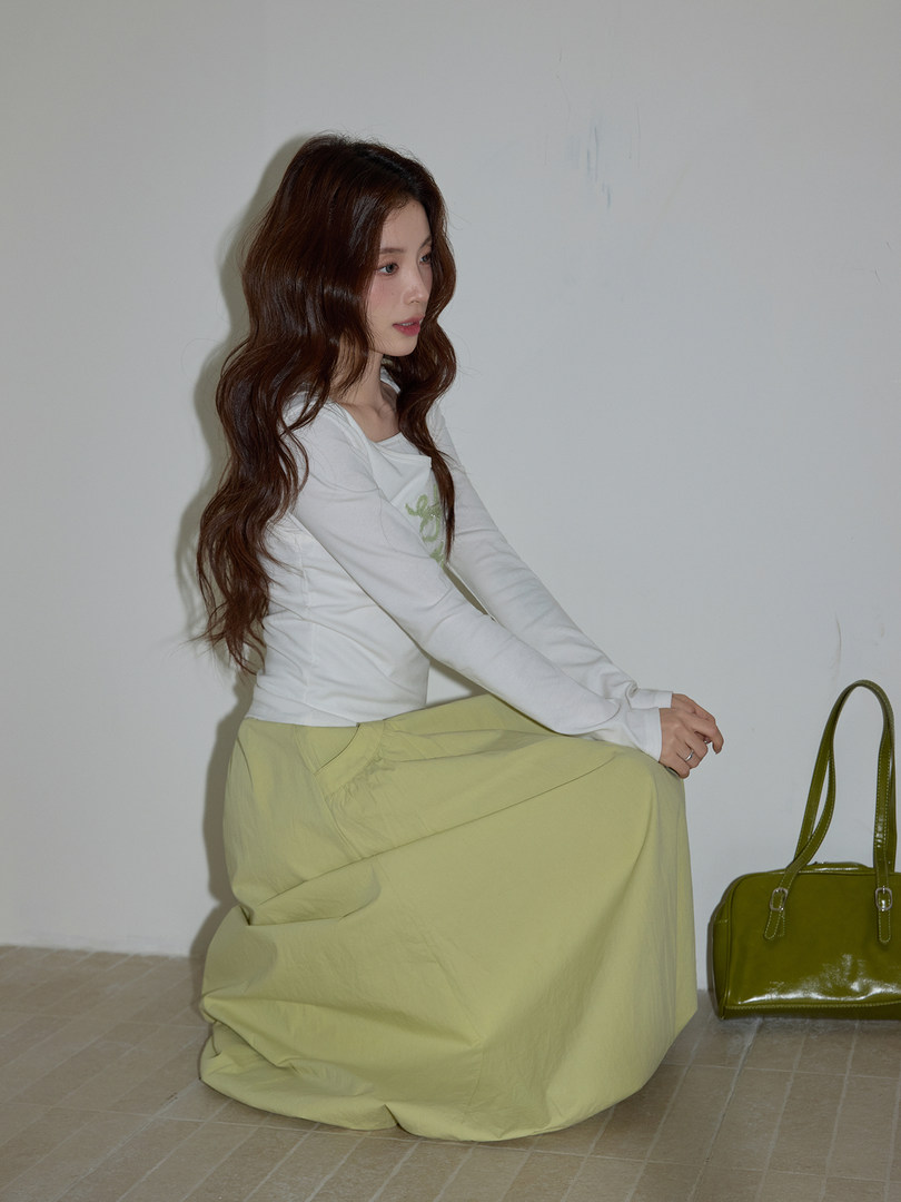 Model wearing a white long-sleeve top with a pale green skirt, styled with a green handbag