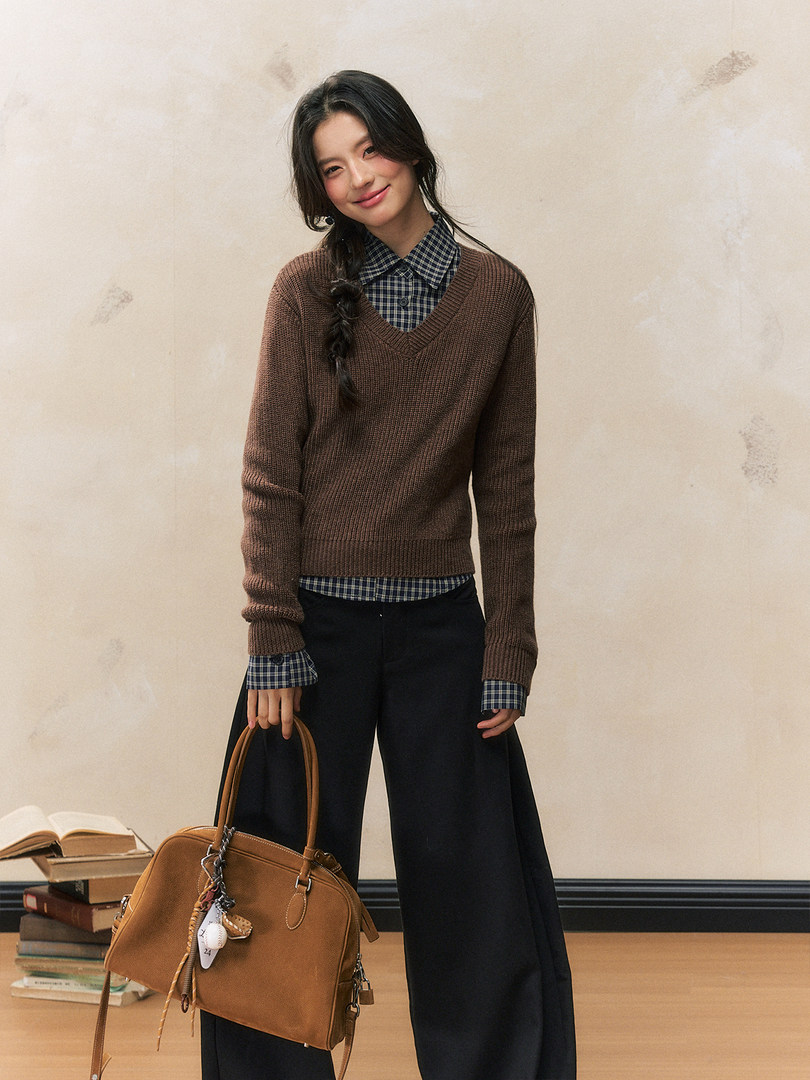Woman wearing a brown knit sweater layered over a plaid shirt with black wide-leg pants and a tan handbag