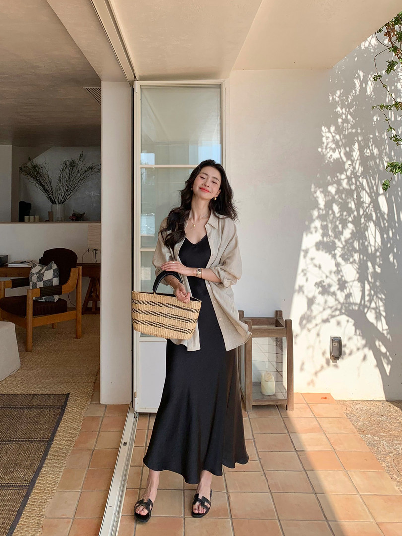 Woman wearing a black dress with a beige overshirt, black sandals, and a woven handbag outdoors