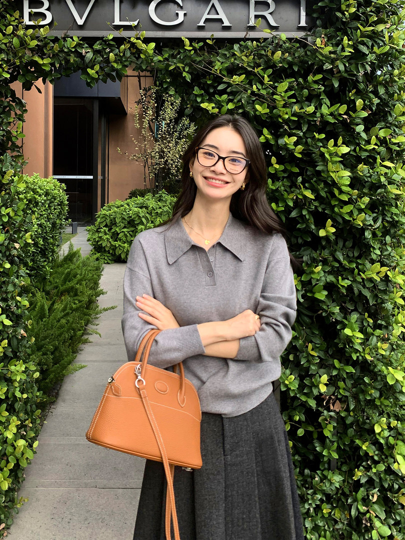 Woman wearing a gray knit top and holding a tan leather handbag outdoors