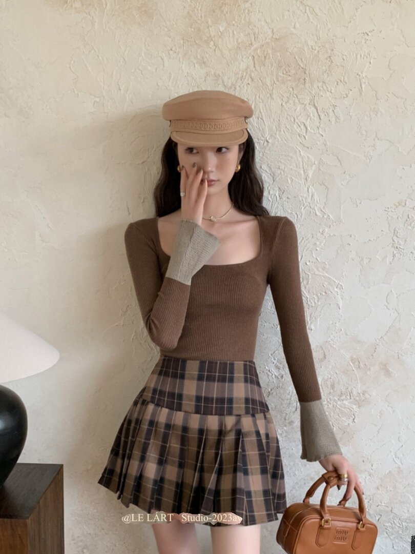 Model wearing a brown long-sleeve top with a plaid pleated skirt and holding a small handbag