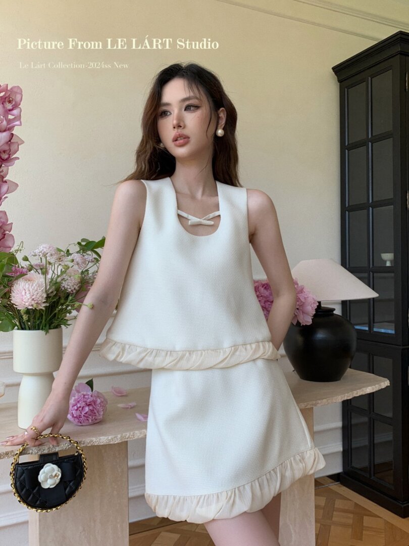 Model wearing a white sleeveless dress with ruffle trim and a small black handbag