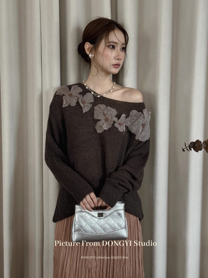 Model wearing a dark brown off-shoulder knit sweater with floral appliques and a silver handbag