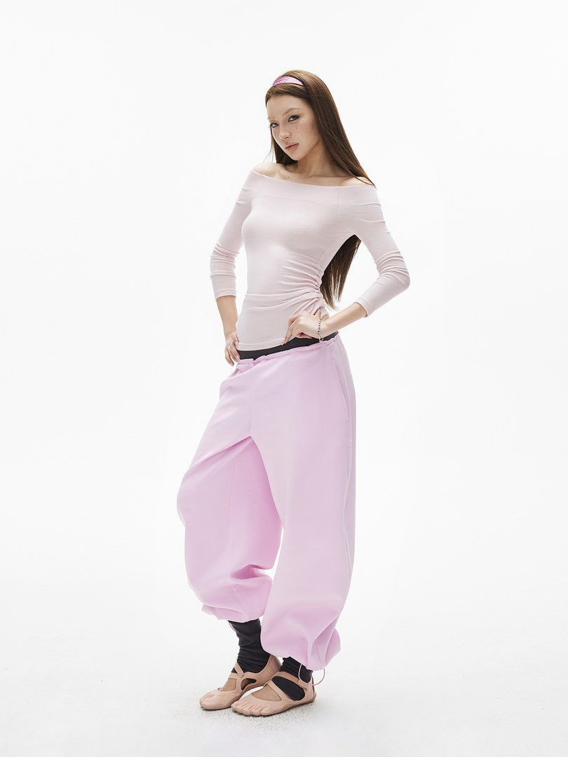 Model wearing a white off-shoulder top with oversized pink pants and beige shoes.