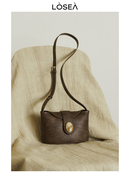 Brown leather shoulder bag with long strap and decorative clasp on a neutral backdrop