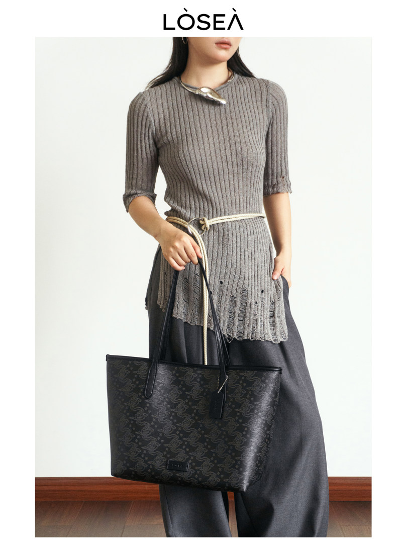 Model wearing a gray ribbed knit top with dark trousers and holding a black tote bag