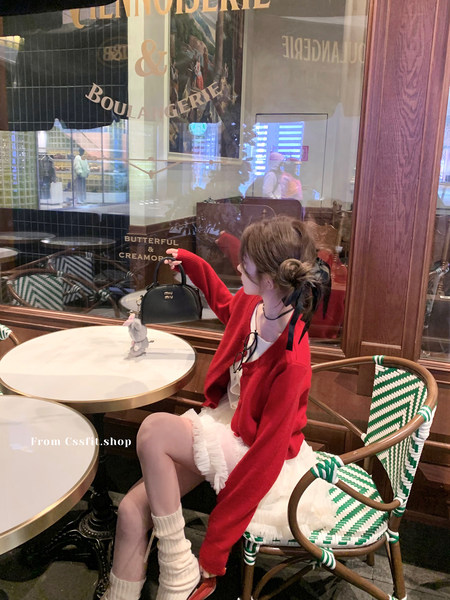 Woman seated in a cafe holding a black Miu Miu handbag while wearing a red cardigan and white skirt