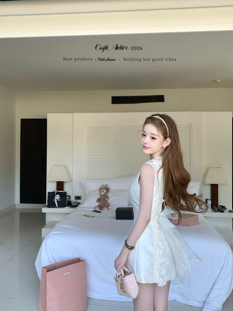 Woman wearing a white lace dress and holding a small pink handbag in a bedroom