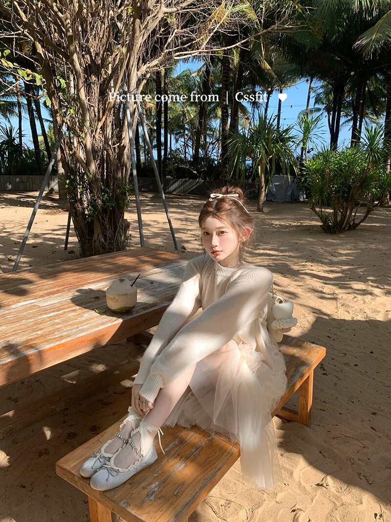 Woman wearing a light cream dress and sweater while sitting on a beach bench