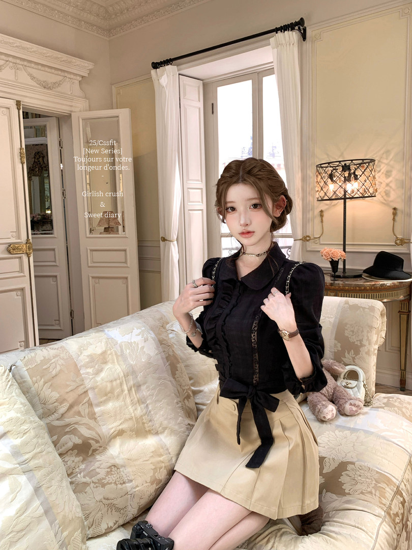 Model wearing a black ruffled blouse with a beige pleated skirt in an indoor fashion photo