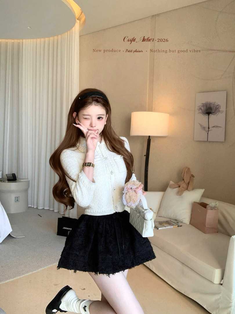 Woman modeling a white textured cardigan with a black skirt and white boots indoors