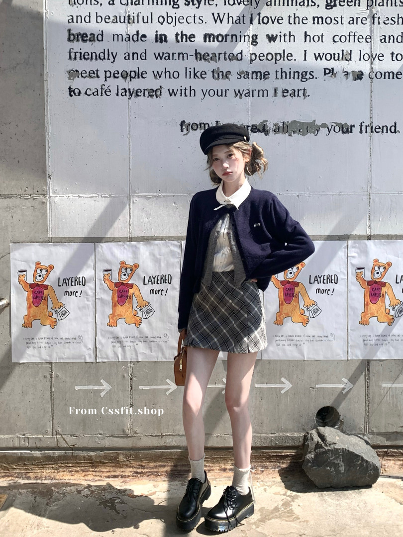 Woman wearing a preppy layered outfit with a black cardigan, plaid skirt, and cap