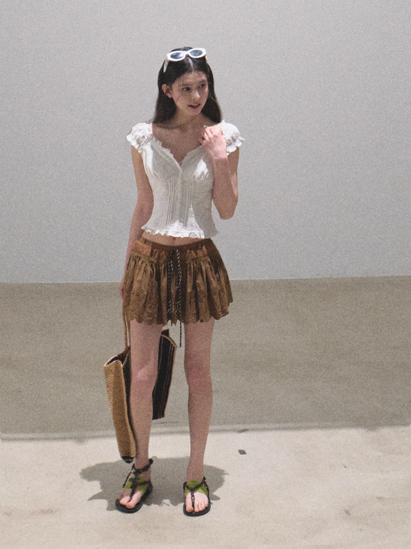 Model wearing a white ruffled top, brown mini skirt, and green-black sandals
