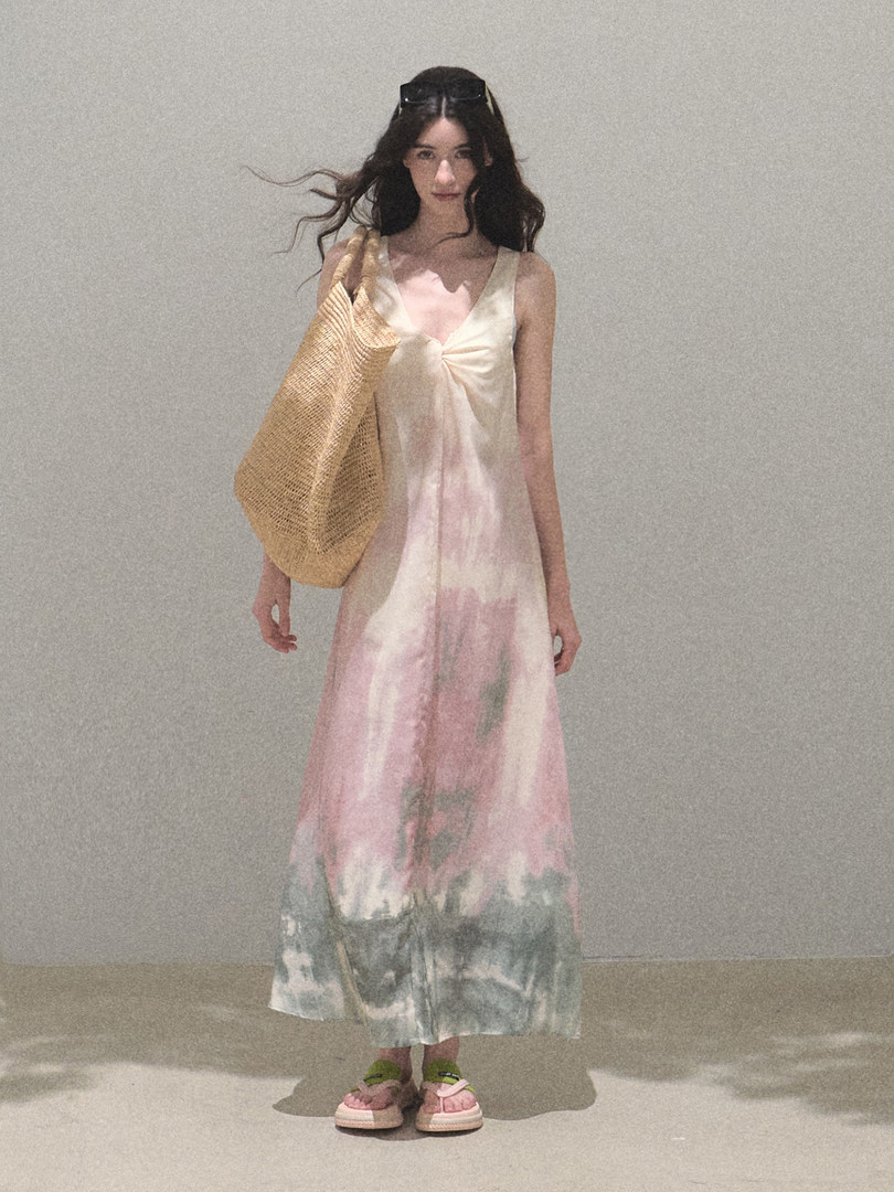 Model wearing a pastel tie-dye sleeveless maxi dress with a woven tote bag and pink sandals