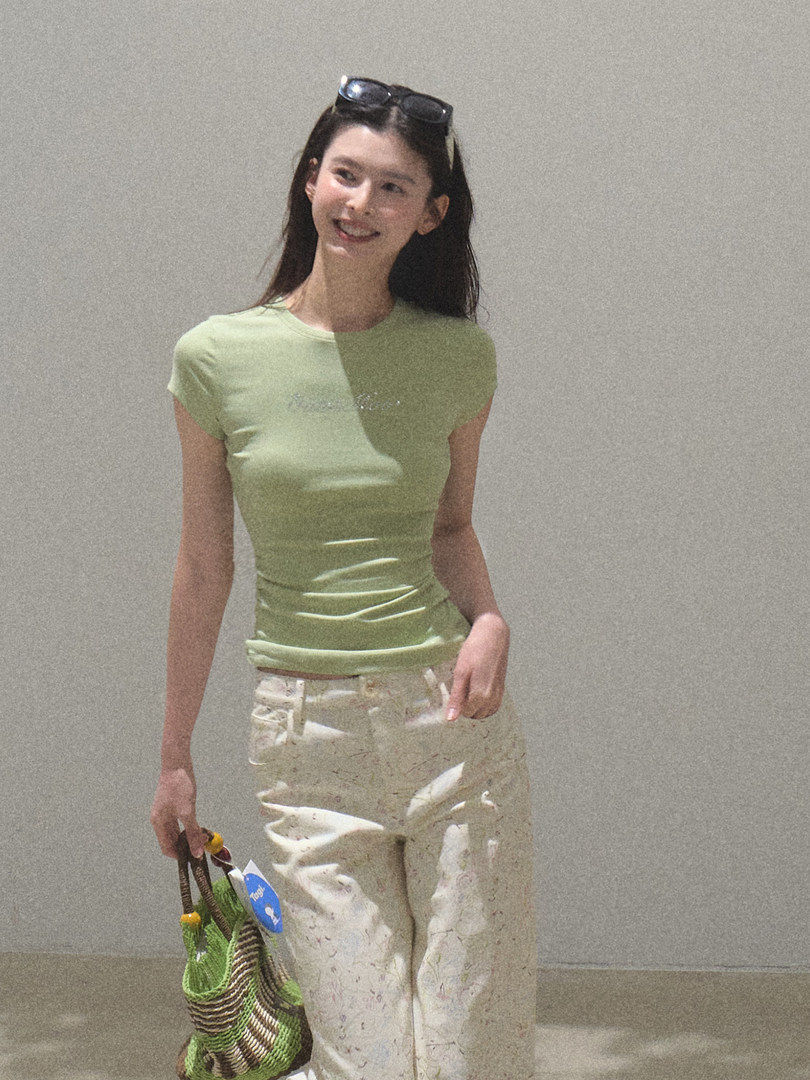 Model wearing a light green fitted T-shirt and cream pants holding a woven bag