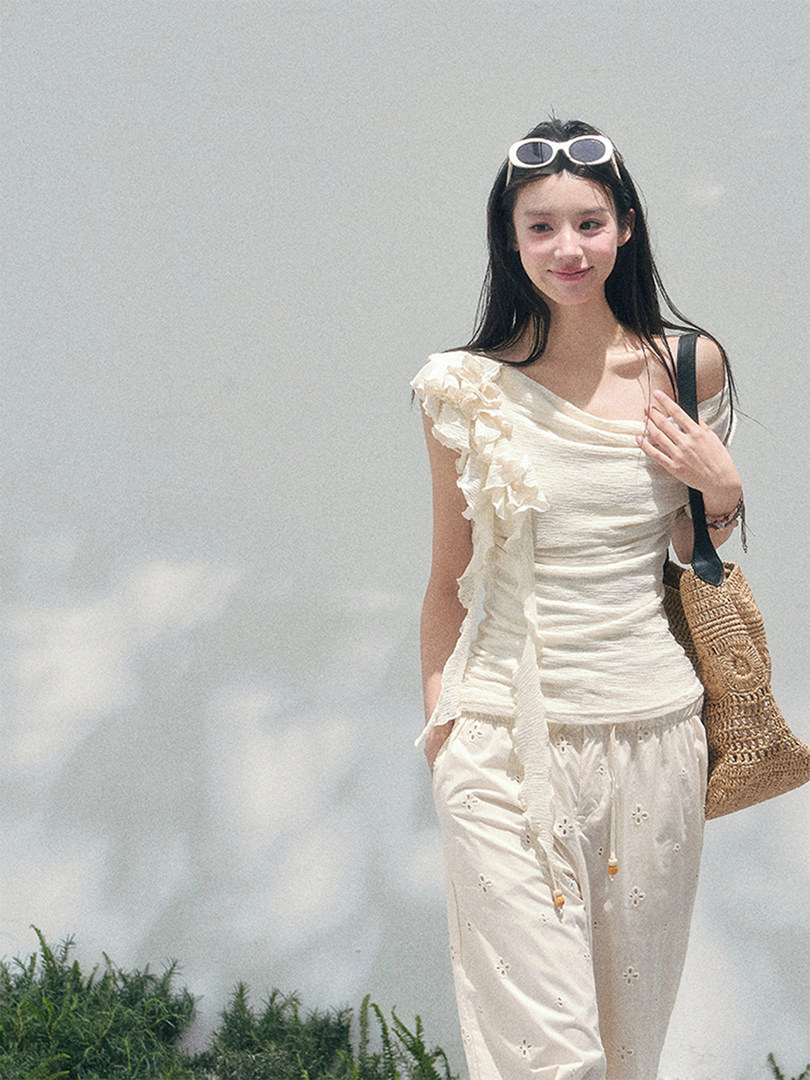 Woman wearing a cream ruffled top and matching white pants with a woven tote bag