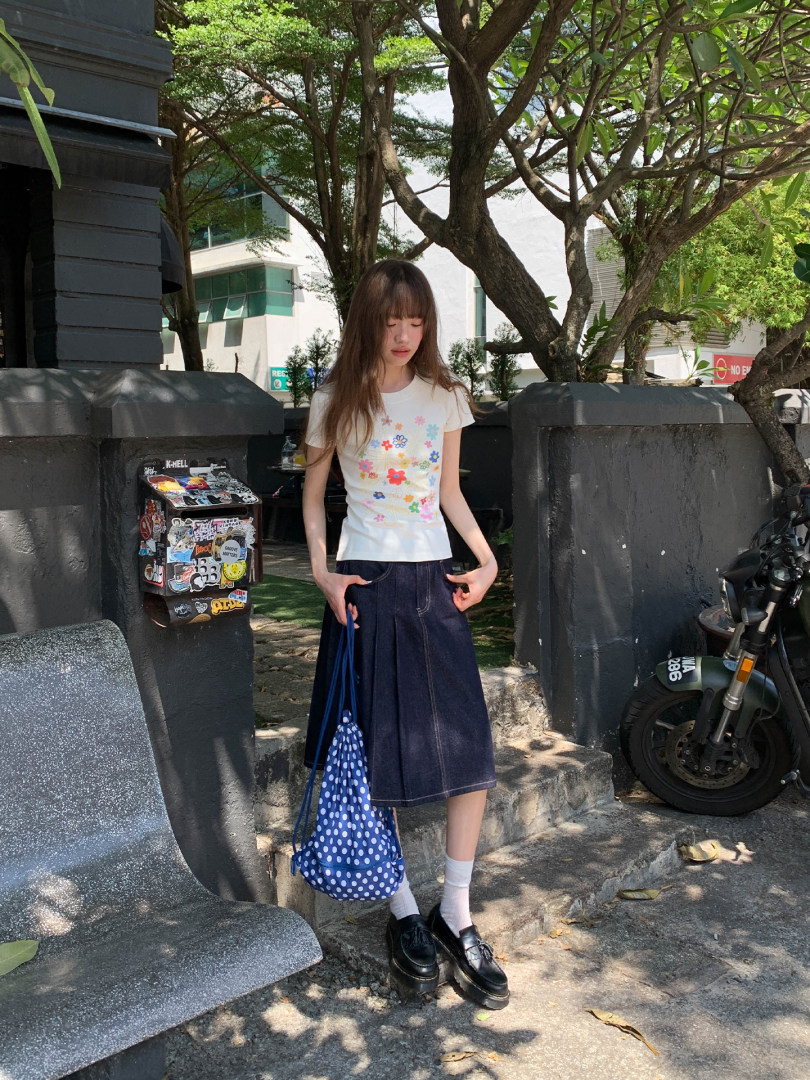 Woman wearing a white floral graphic T-shirt, dark denim skirt, black loafers, and a blue polka dot bag outdoors