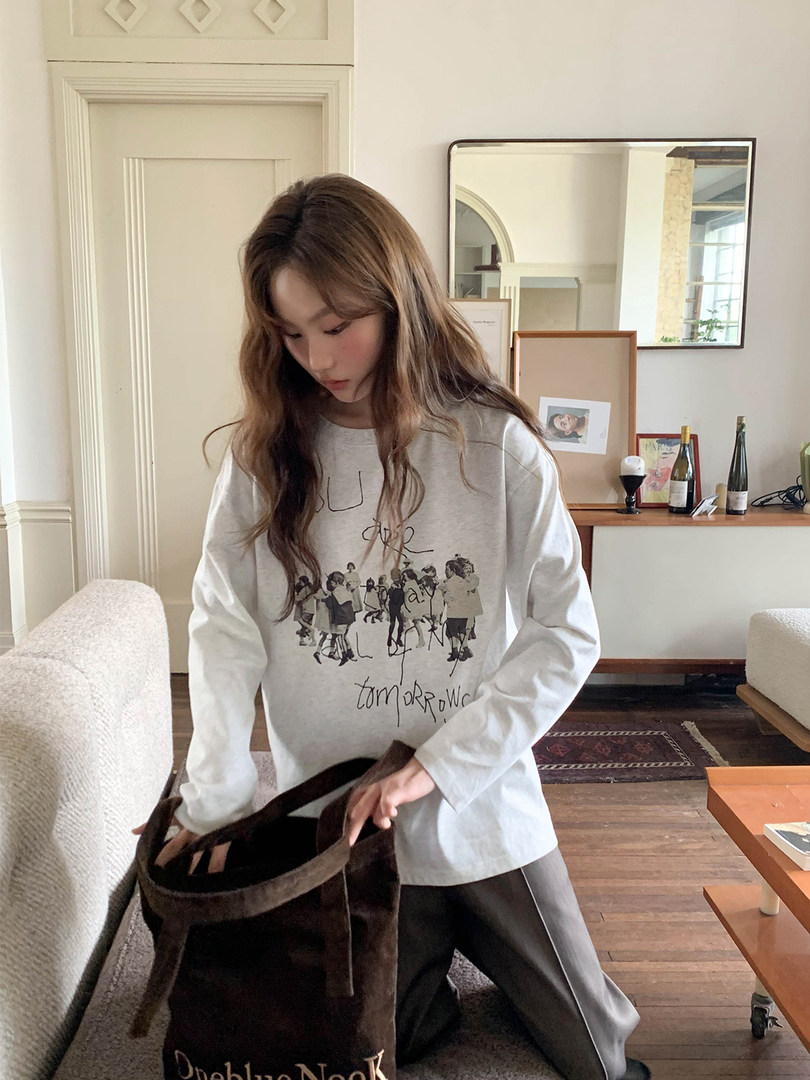 Woman wearing a white graphic long-sleeve top and holding a dark tote bag indoors