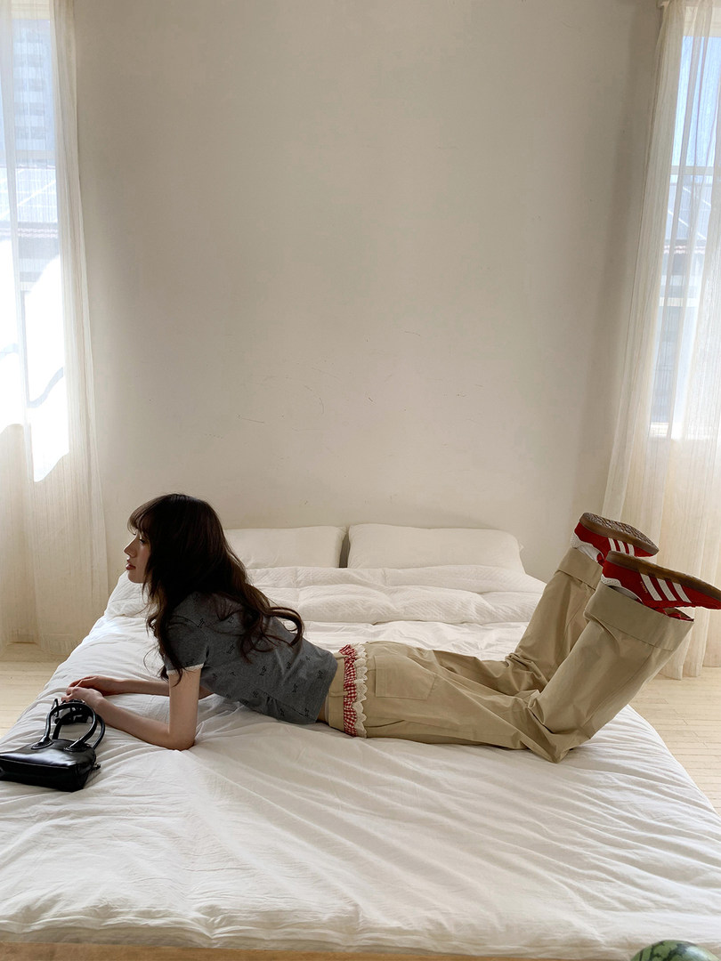 Model wearing a gray crop top and beige pants on a bed with a black handbag