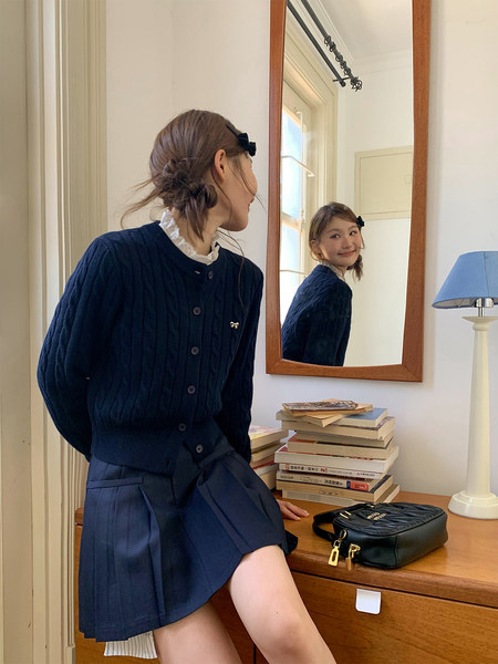 Model wearing a navy knit cardigan and pleated skirt with a black handbag on a dresser