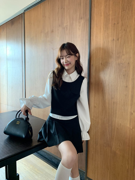 Woman modeling a black sleeveless dress layered over a white blouse, holding a black handbag.